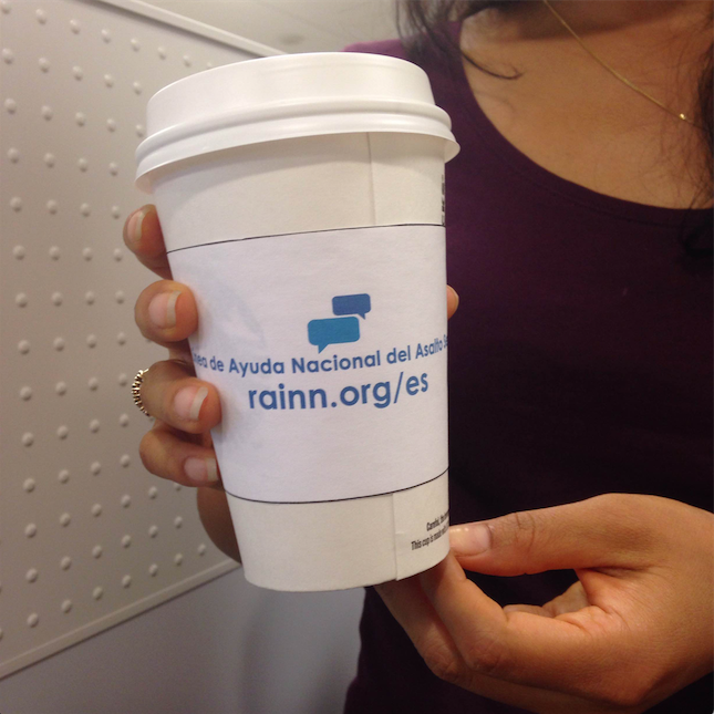 Person holds a coffee cup with a sleeve. On the sleeve is written "Linea de Ayuda Nacional del Asalto Sexual" and "rainn.org/es" 